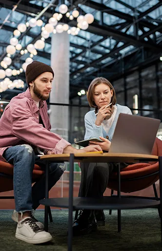 Two coworkers sit at a low table in a modern office, looking at a laptop together