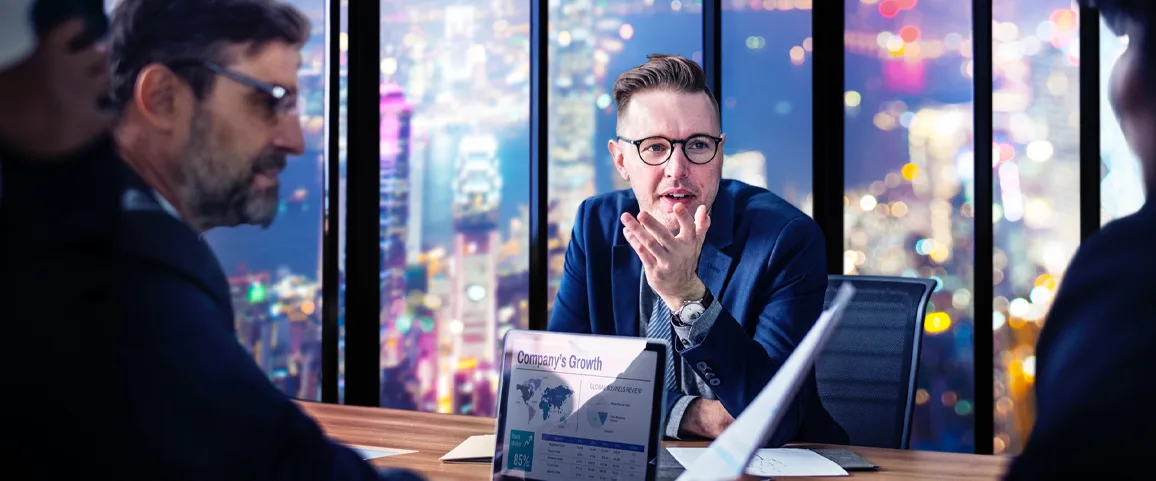 Businessman in a blue suit and glasses speaks to colleagues at a night-time city office; laptop shows growth charts titled 'Company's Growth'.