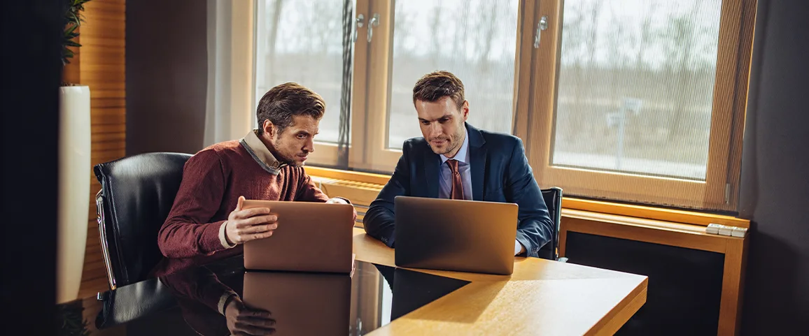 Two business colleagues discuss at a wooden table, each using a laptop in a sunlit office.]','Two men in smart casual and a suit work together on laptops at a conference table.]','Co-workers collaborating on a project in a modern office with large windows.]','Two professionals reviewing data on their laptops during a meeting at a glass-topped table.]','Business partners discussing strategy at a table by windows in a bright office.