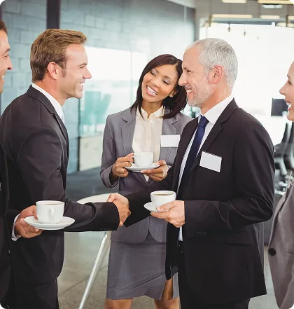 Group of four professionals in business attire chatting and holding coffee cups in a modern office lobby setting for a networking break.