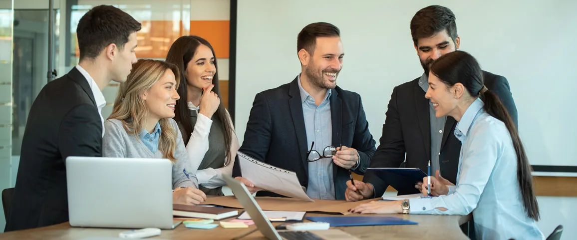 A group of professionals collaborating around a table, smiling as they review documents and laptops in a modern office.