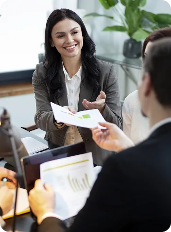 Smiling businesswoman in a gray blazer hands a chart to colleagues during a meeting in a bright office.