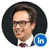 Professional headshot of a smiling man with dark hair and glasses, wearing a suit and red tie against a dark background, suitable for a profile image.