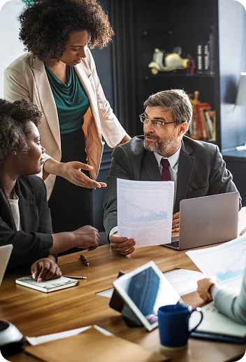 Team of professionals at a conference table; a man in a suit holds a document as colleagues discuss.