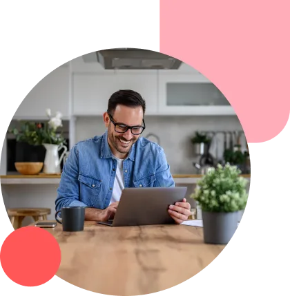 Man wearing glasses and a denim shirt uses a tablet at a wooden kitchen table, smiling, with a mug and potted plant nearby.