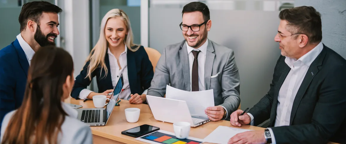 Five business professionals sit around a conference table, smiling and discussing documents and laptops in a bright office.