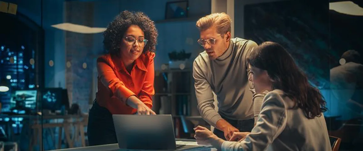 Three coworkers gather around a laptop on a desk, collaborating during a nighttime office meeting.
