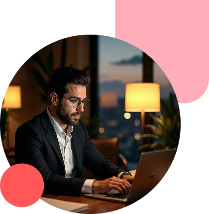 Man in a suit wearing glasses types on a laptop at a desk in a dimly lit office, with a city skyline visible through a window in the background.