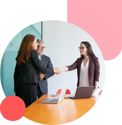 Three professionals shake hands over a conference table with laptops in a bright office setting.