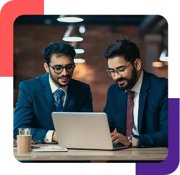 Two business colleagues in suits collaborating over a laptop at a desk in a modern office setting.