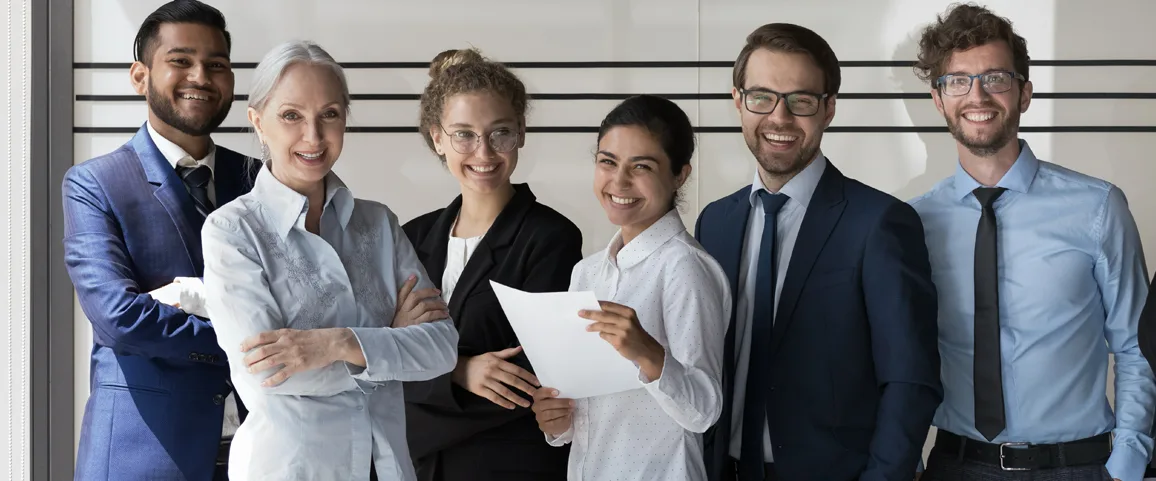 Six professionals in business attire smiling together in an office, one holding a sheet of paper as they pose for a group photo.
