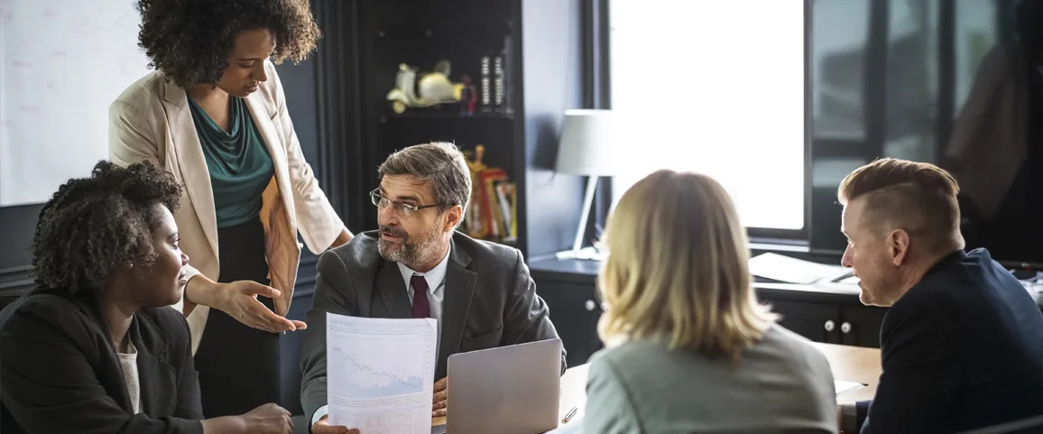 Group of five professionals in a meeting around a conference table; a woman stands and explains a document to a seated man in a suit holding a paper and laptop.