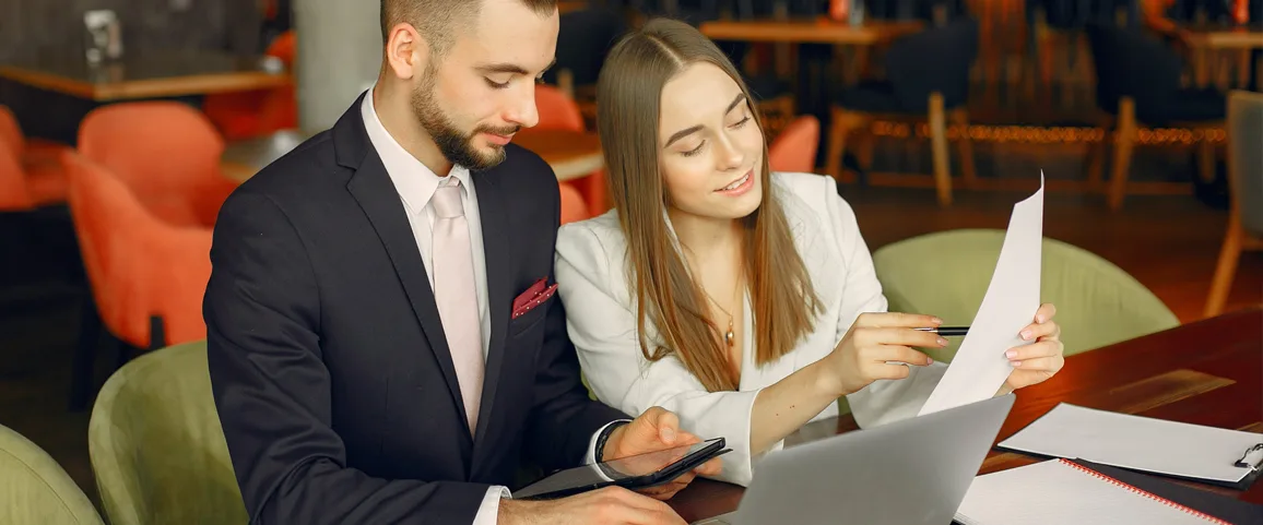 Two business colleagues in a suit and white blazer review documents at a wooden table with a laptop and tablet in a casual office cafe setting.