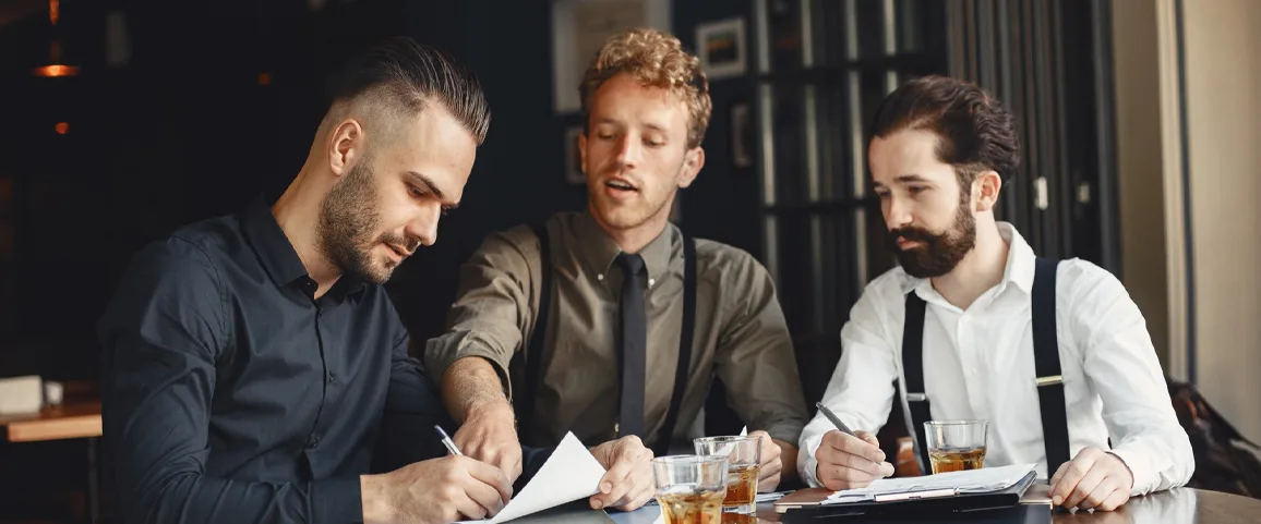 Three colleagues in a casual office setting review documents at a table, writing notes as they discuss.