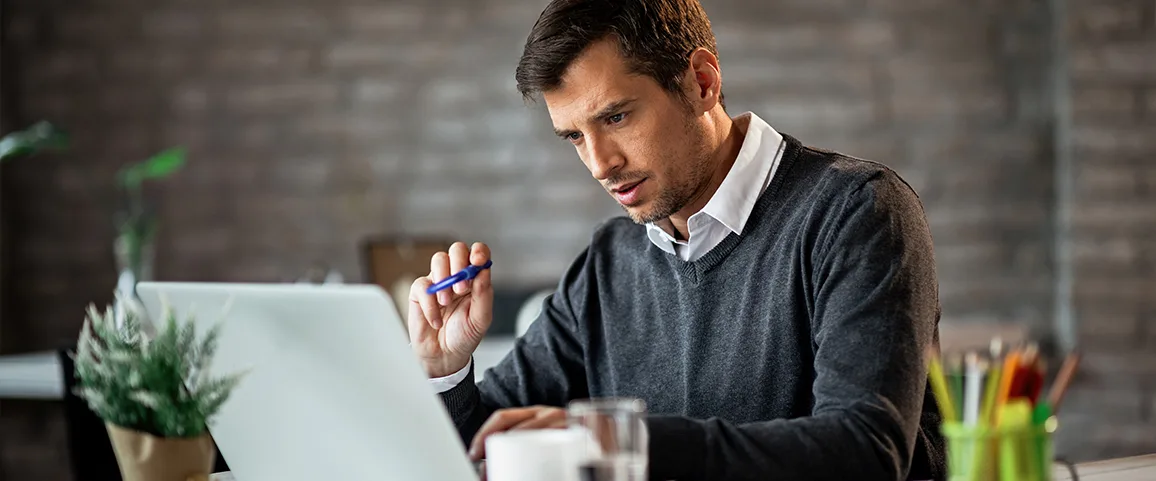 Man in a gray sweater works on a laptop at a desk, holding a blue pen and looking at the screen.