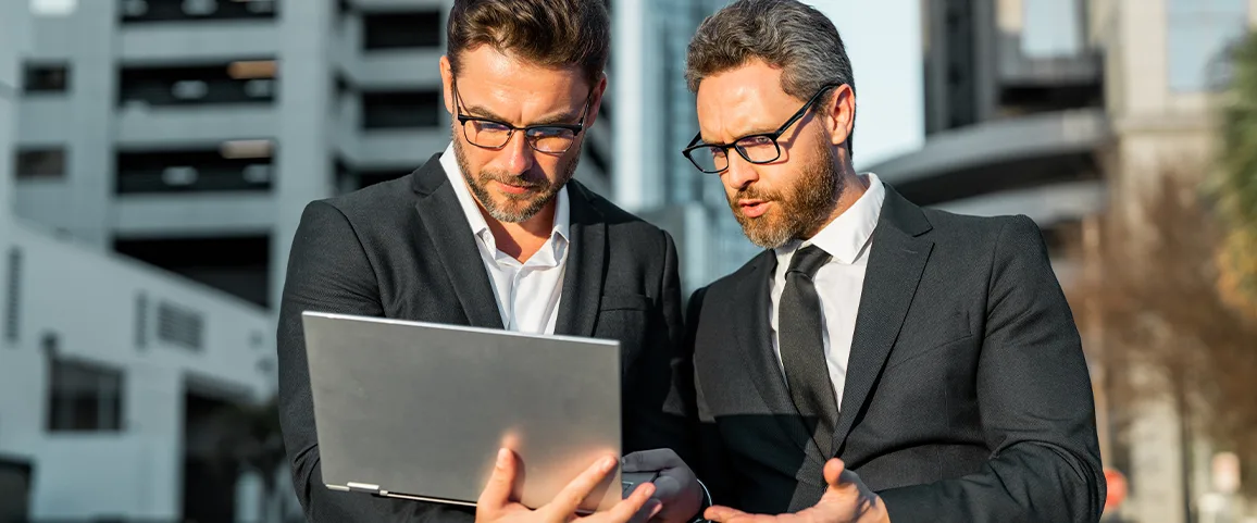 Two suited men with glasses review a laptop outdoors in an urban setting.