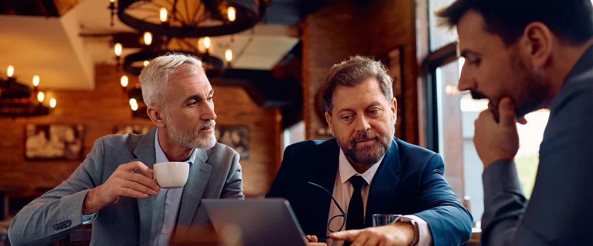 Three businessmen in a brick-walled cafe discuss a tablet, one holding a coffee cup.