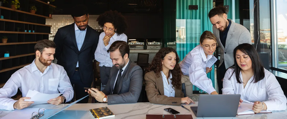 Group of eight professionals around a conference table collaborating on documents and a laptop in a modern office, sharing ideas.