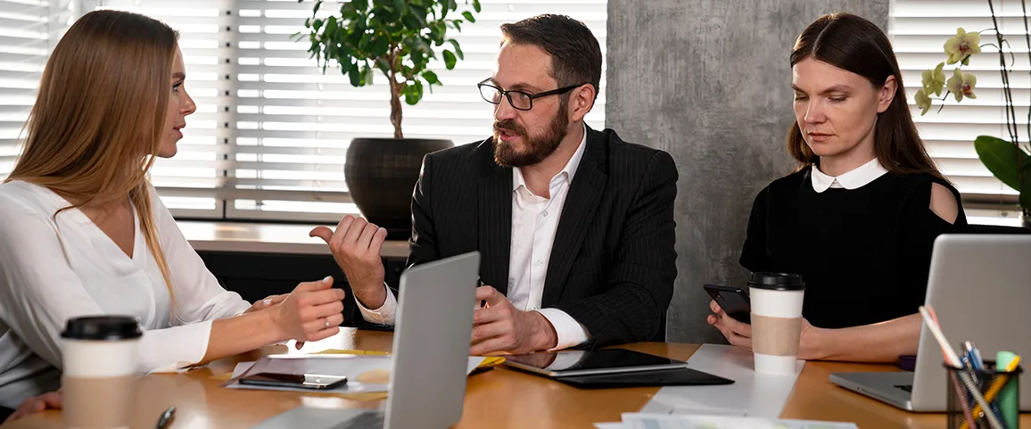 Three coworkers in a modern office discuss a project around a table with laptops, papers, and coffee cups on it