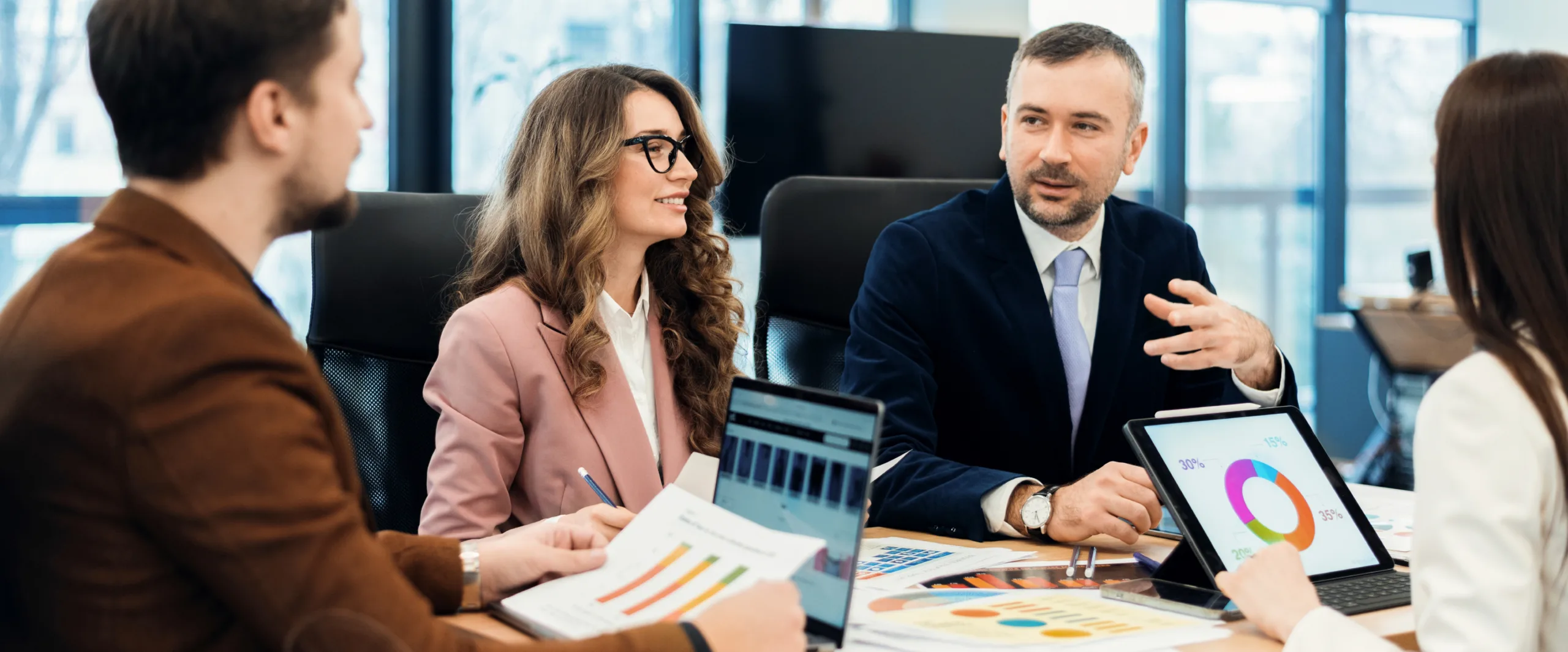 Team of professionals in a meeting discussing charts on tablets and printed graphs at a conference table in a bright office setting.
