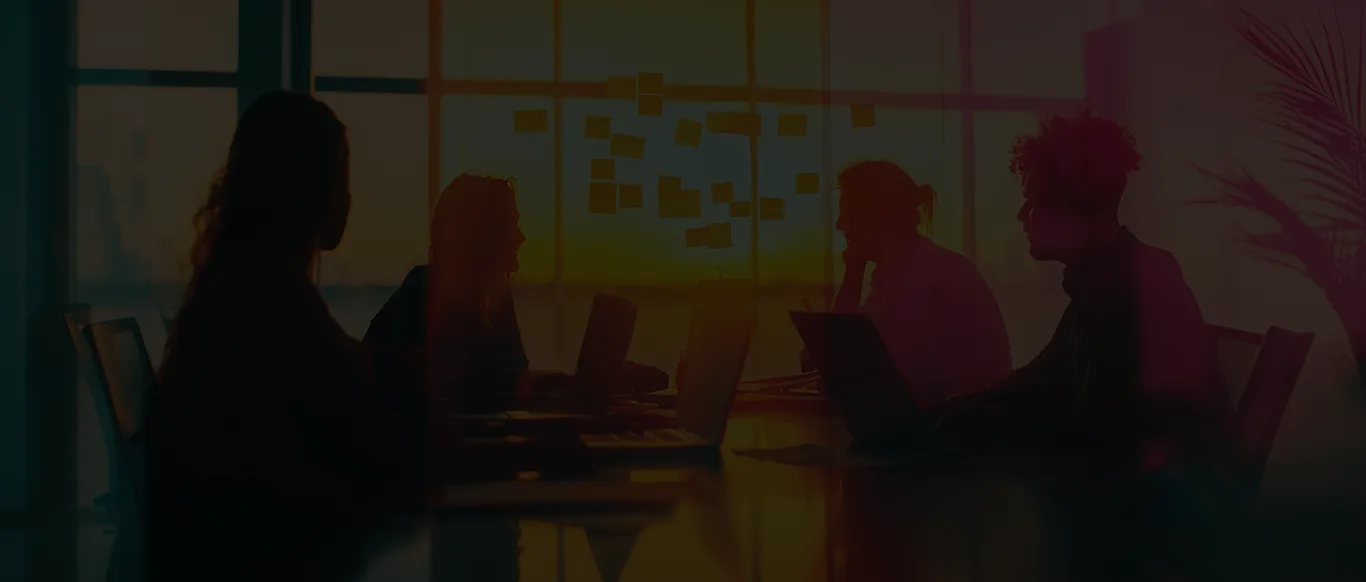 Team of four in a meeting room brainstorming, with sticky notes on a glass wall and laptops, backlit by sunset light.