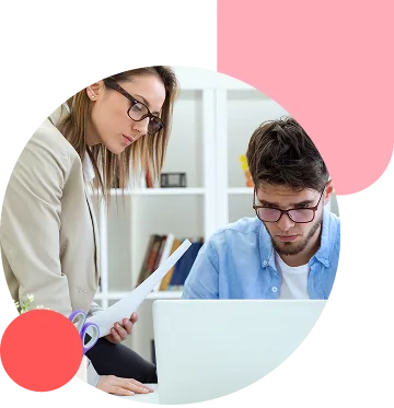 Two colleagues collaborate at a desk: a woman stands with documents while a man sits, both wearing glasses in a bright office.