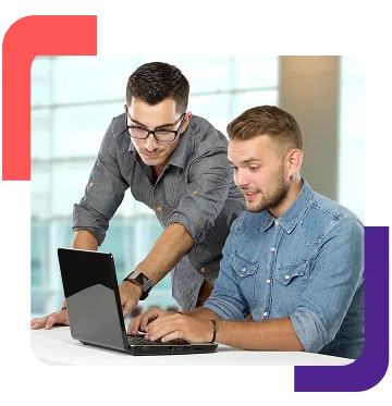 Two colleagues collaborate at a laptop on a white table in a bright office setting, one standing and pointing at the screen.