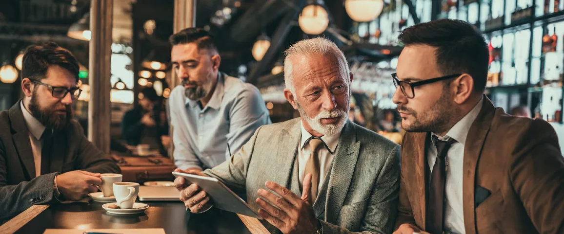 Group of four professionals in a cafe, one showing a tablet to the others during a meeting over coffee.