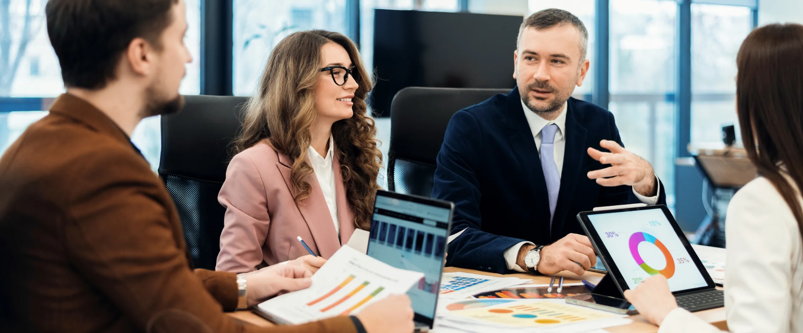 Team of professionals in a modern office discussing colorful charts and laptops around a conference table, mid-meeting.