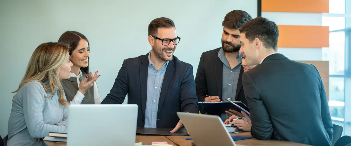 Group of five coworkers in business attire collaborating around a table with laptops and documents, smiling and talking in a bright office.