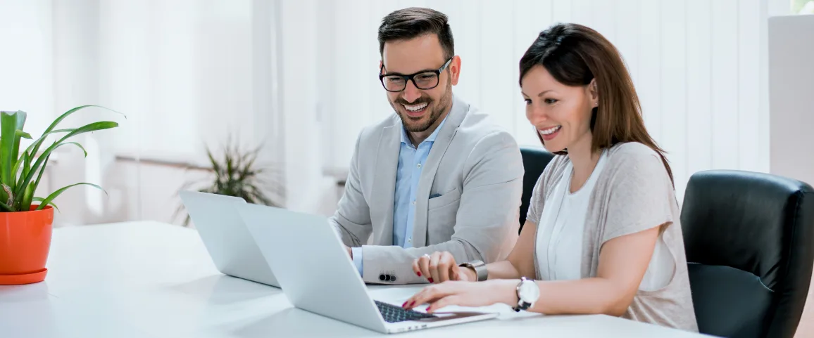 Two coworkers collaborate at a white desk with an open laptop in a bright office setting, smiling at the screen.