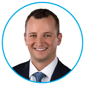 Professional headshot of a smiling man in a navy suit and blue tie against a white background, framed in a blue circular border.