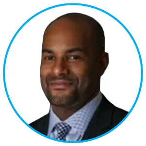Professional headshot of a bald man with a trimmed beard, wearing a dark suit and light blue patterned tie, against a white background.